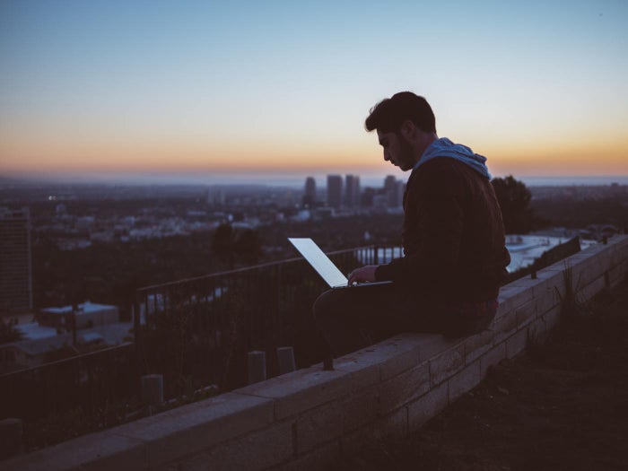 Photo of man using a computer sitting on a hill overlooking a city at sunset.